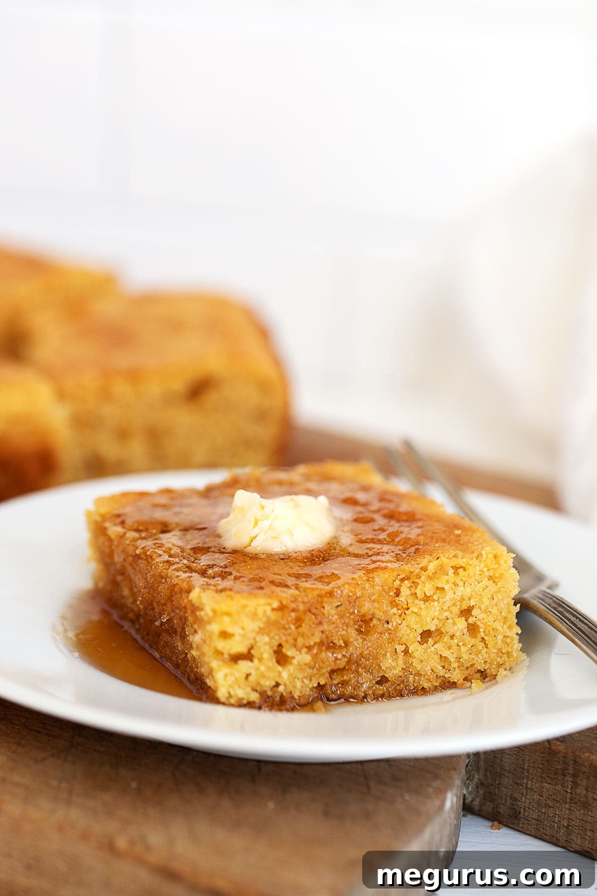 Warm Johnny Cake slices, glistening with melted butter and sweet maple syrup, served on a white plate with fresh spring flowers in the background.