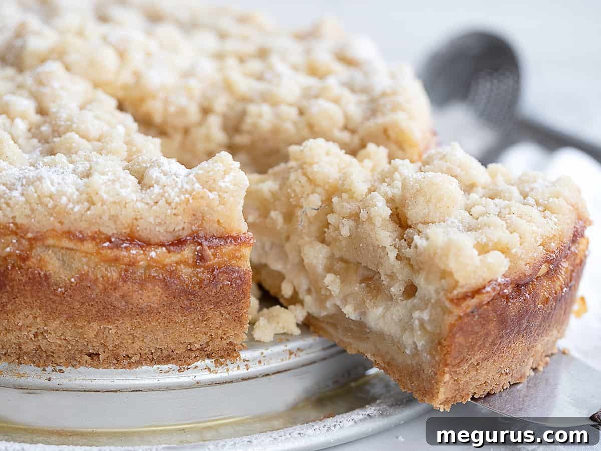 A slice of delicious apple crumb cake on a white plate, showcasing its distinct layers of crust, apples, creamy filling, and crispy crumb topping.