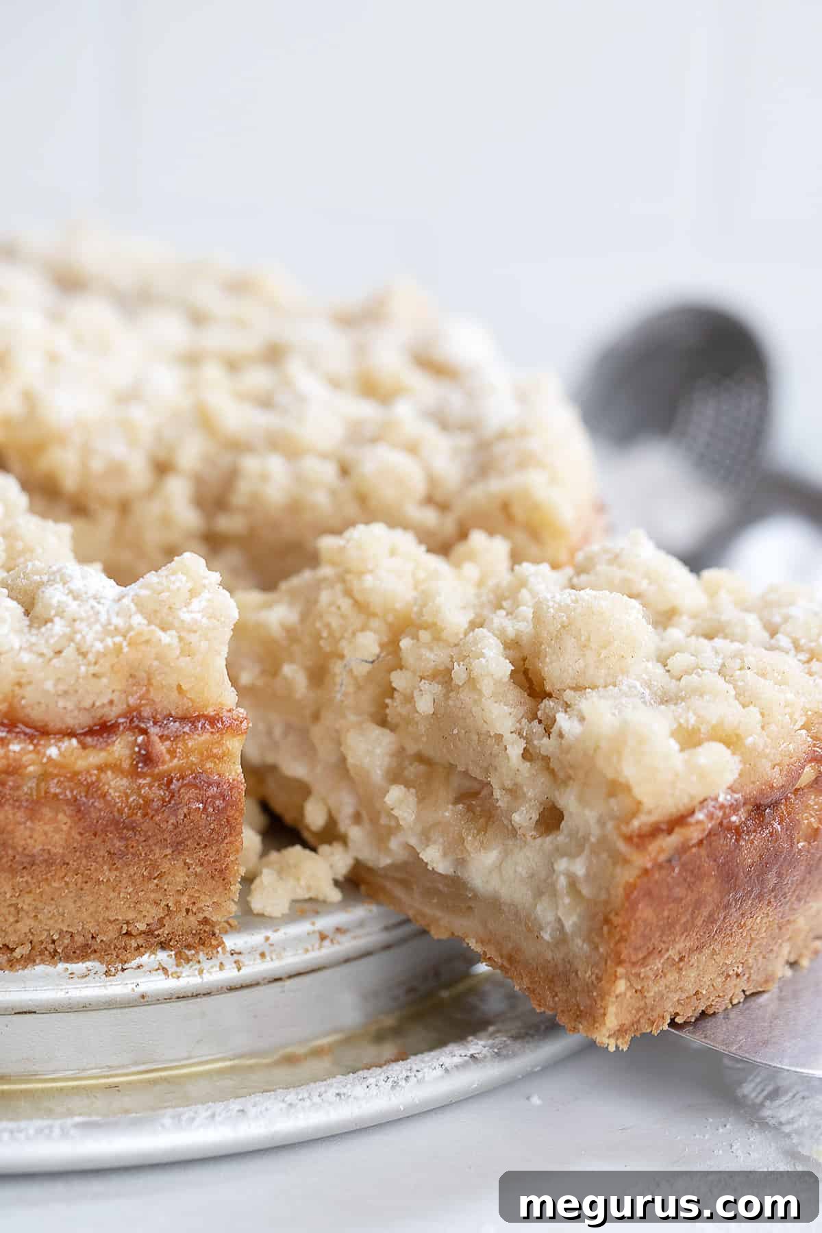 A close-up shot of a perfect slice of apple crumb cake on a plate, highlighting the crispy golden crumble topping and the creamy apple layers beneath.