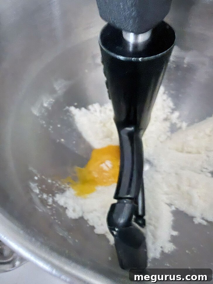 A single egg yolk being added to the butter and sugar mixture in a stand mixer bowl, ready to be incorporated.