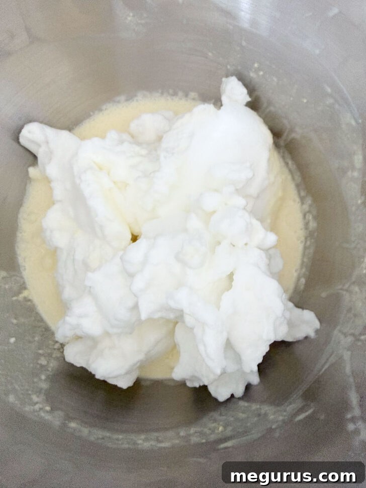 Fluffy whipped egg whites gently spooned over the lemon batter in a mixing bowl, prior to being folded in.