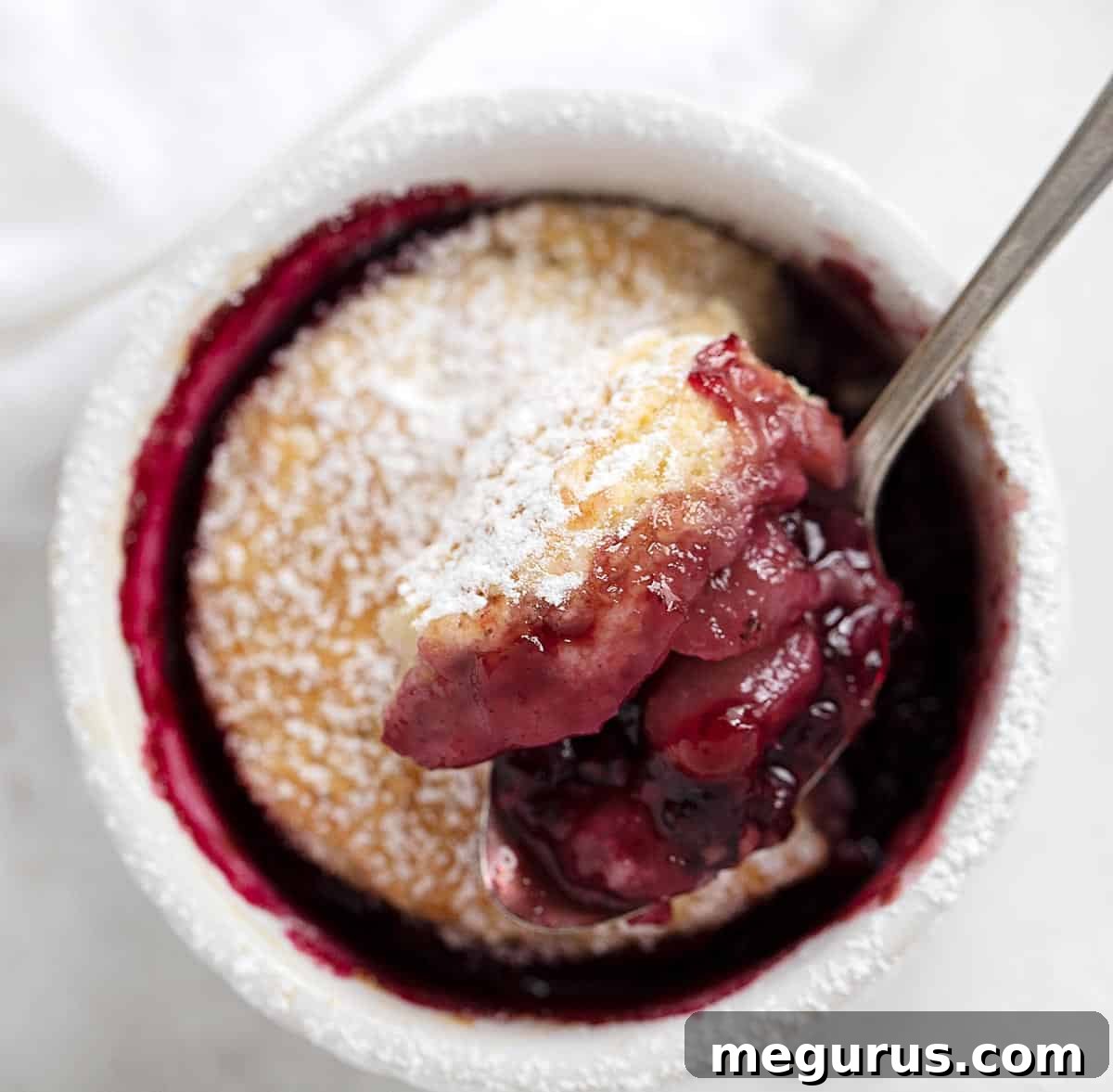 A freshly baked blackberry pear cobbler in a ramekin, with a spoon inserted, showing the bubbly fruit and golden topping.