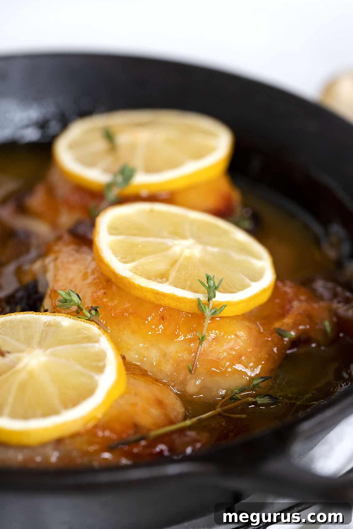 Close-up of glazed lemon chicken thighs in a cast iron pan, garnished with fresh herbs and lemon slices.