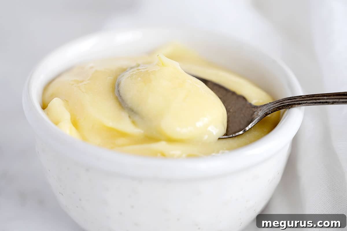 Freshly made lemon curd in a small white bowl with a spoon, garnished with lemon zest.