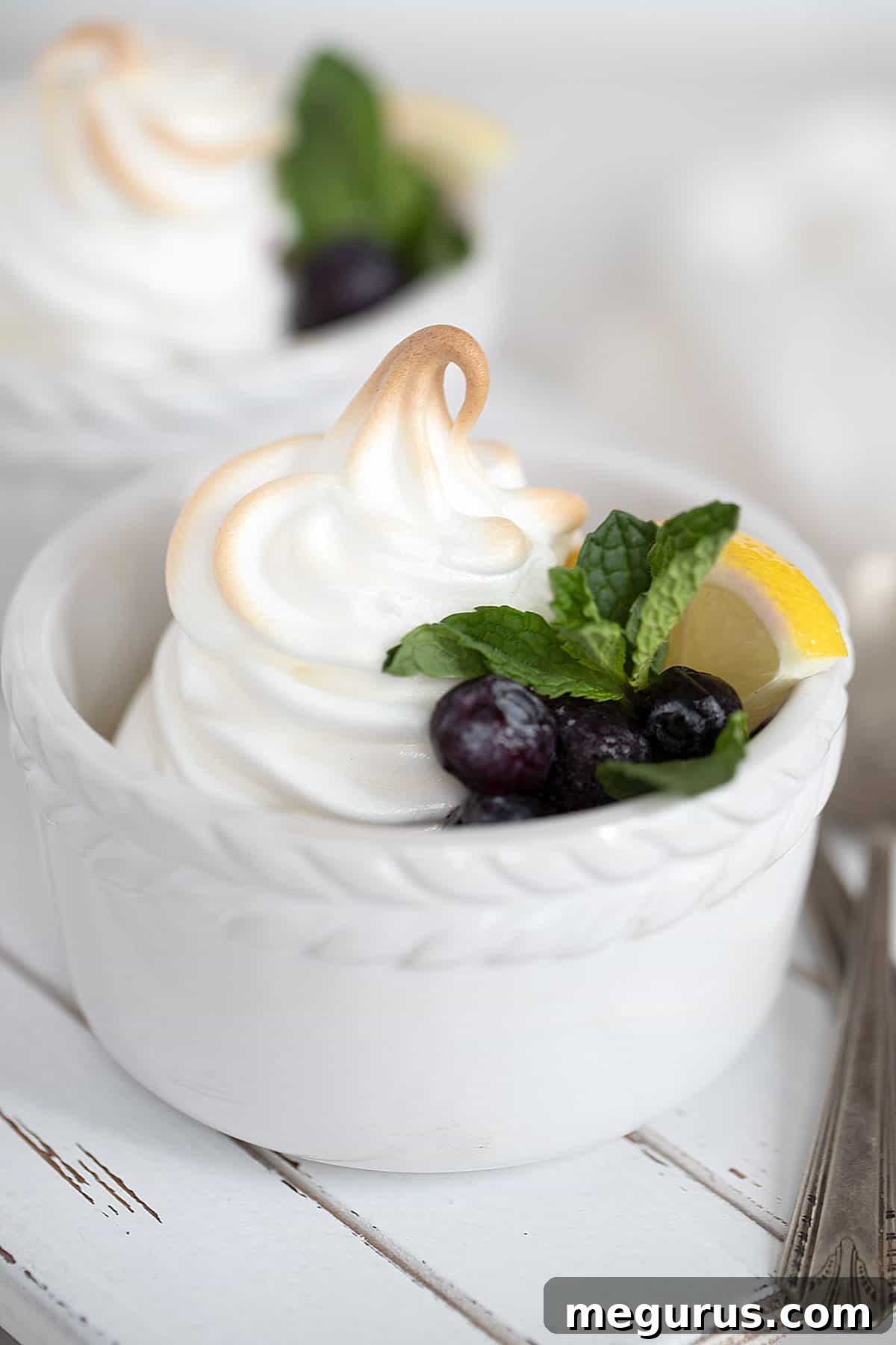 Close-up of baked lemon meringue pudding in a white ramekin, topped with golden meringue and fresh blueberries.