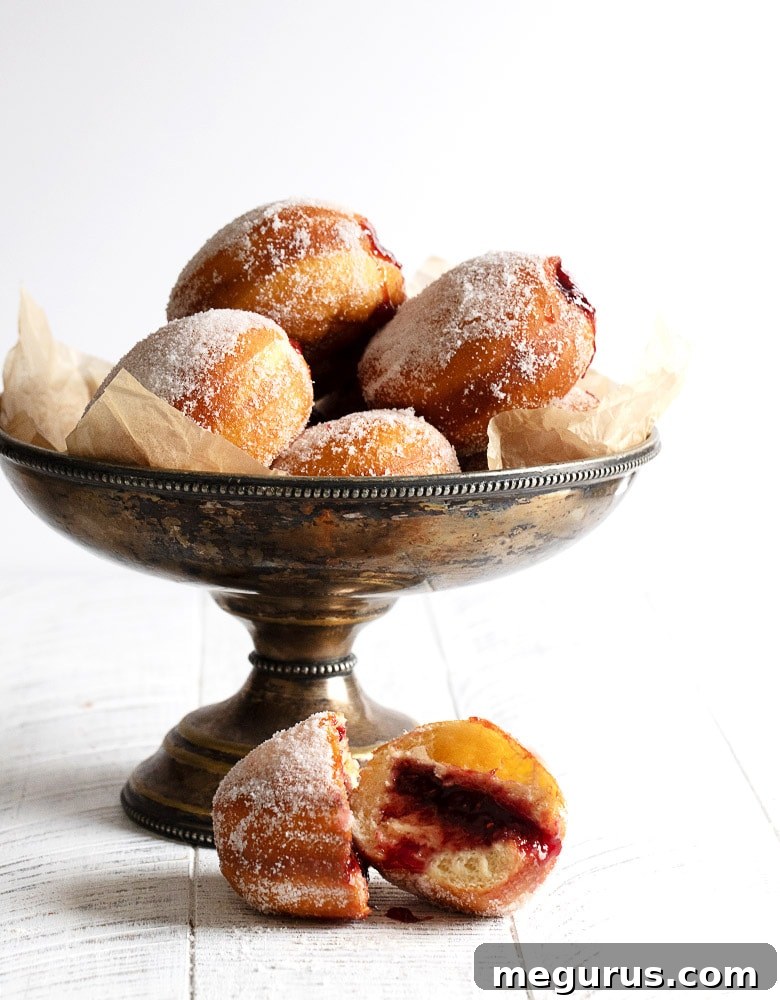 A silver bowl filled with jam-filled paczki, with one cut open to reveal a vibrant raspberry filling.