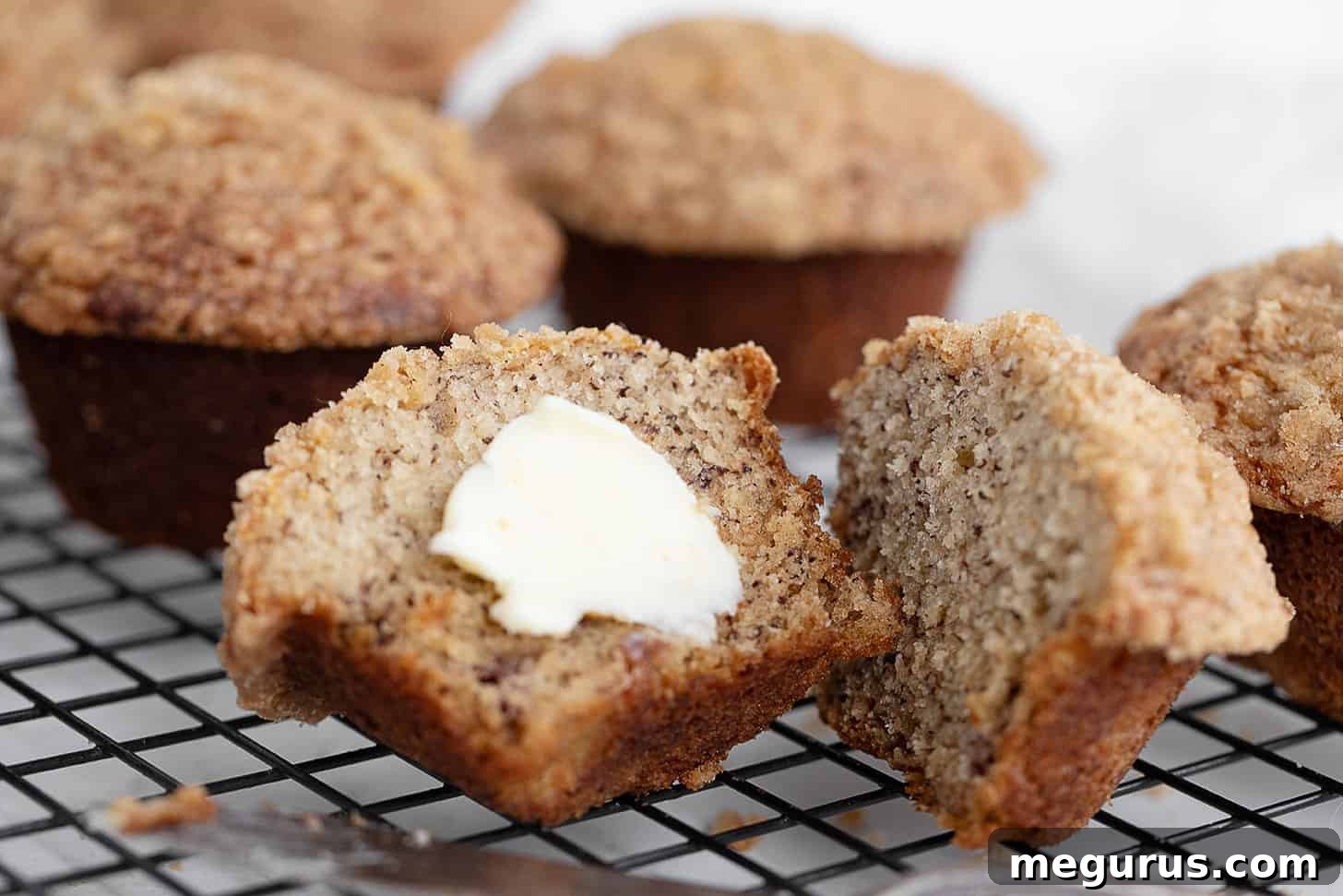 Golden brown banana crumb muffins cooling on a wire rack, with one muffin split open to reveal its tender interior and a dollop of butter.