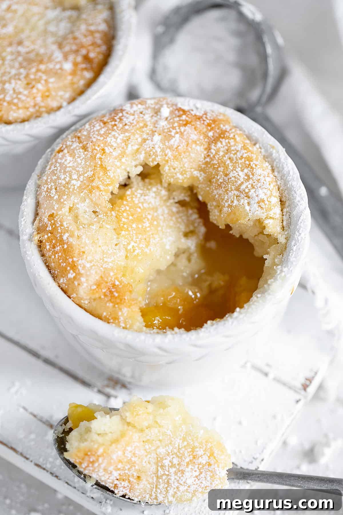 Close-up of a peach pudding cake in a white ramekin, garnished with a dusting of powdered sugar and a spoon.