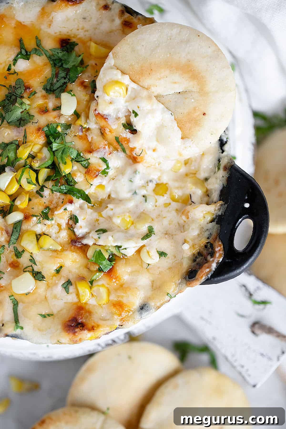 Glorious golden-brown hot corn dip served in a cast iron pan with a side of perfectly baked pita rounds and a garnish of cilantro and chili oil.