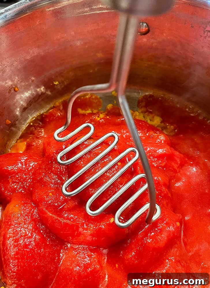 Whole canned tomatoes being mashed directly in the pot with a potato masher, releasing their juices.