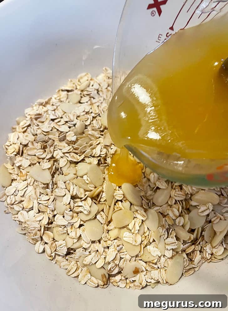 A stream of honey mixture being poured over toasted oats and almonds in a large mixing bowl.
