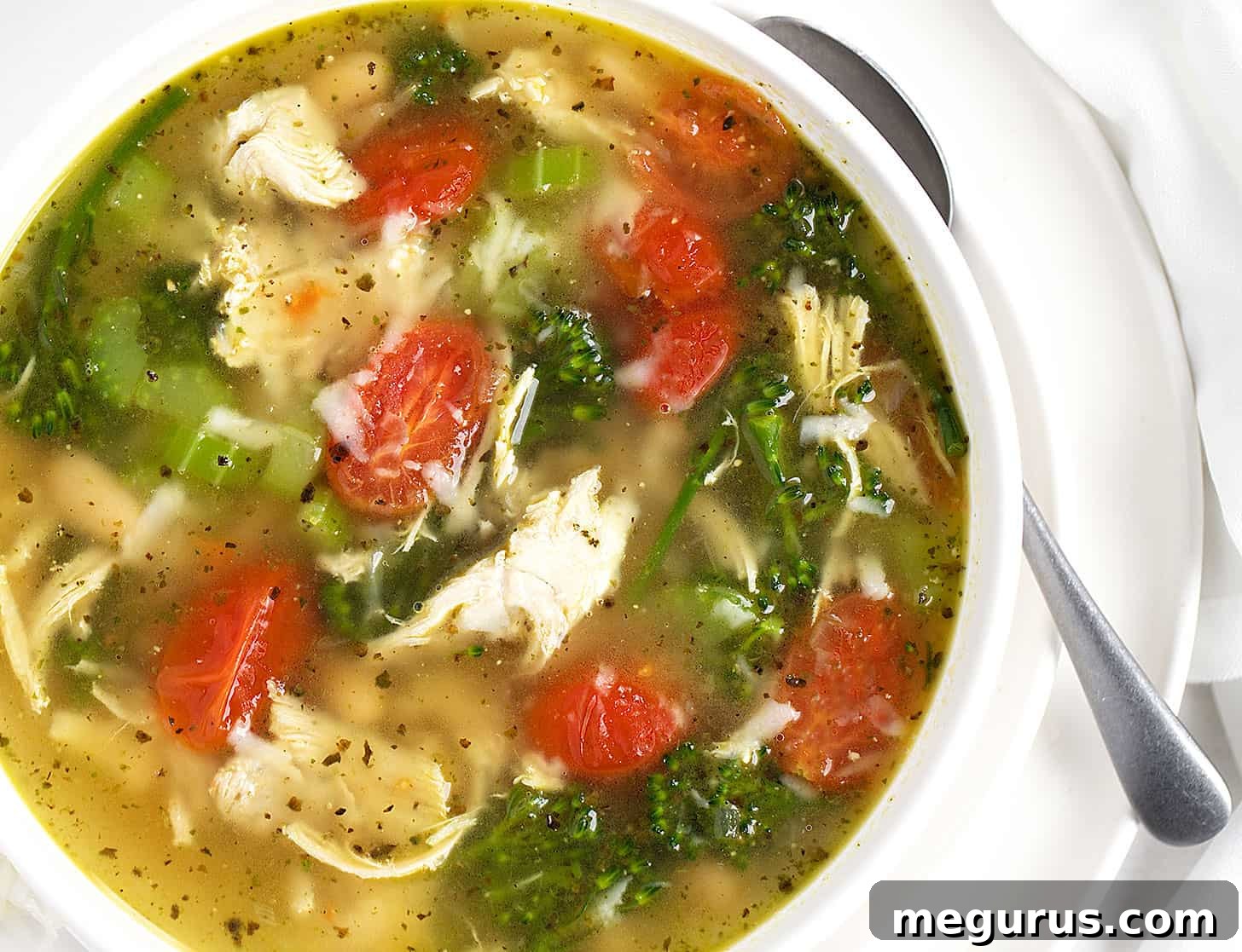 A hearty bowl of Chicken Pesto Soup garnished with fresh basil leaves, featuring shredded chicken, cherry tomatoes, and broccolini.