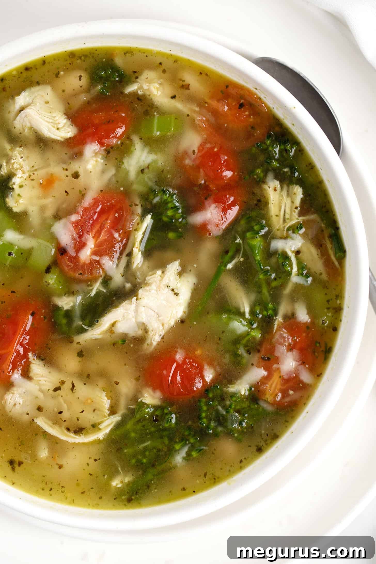 Close-up of a white bowl filled with creamy Chicken Pesto Soup, showcasing shredded chicken, colorful vegetables, and a spoon ready to dive in.