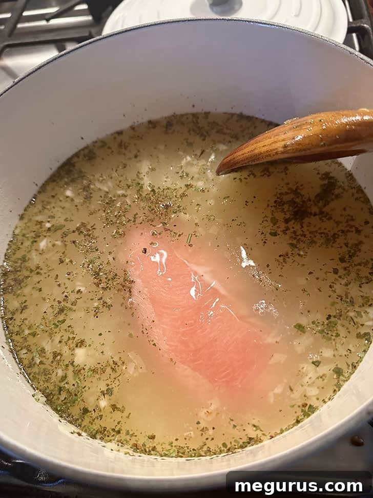 Gently placing a boneless chicken breast into the simmering seasoned broth for poaching.