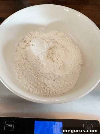 Measuring cup with flour being weighed on a kitchen scale, alongside other dry ingredients.