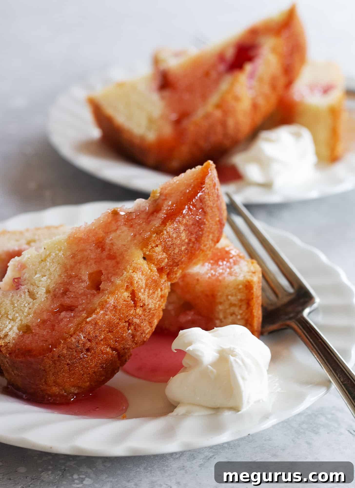 Slice of rhubarb loaf cake on a white plate with a fork and a dollop of whipped cream, showcasing its moist texture.