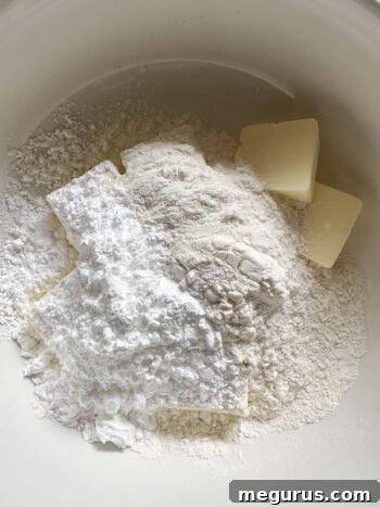 Bowl with ingredients for shortbread base: flour, cold butter pieces, and confectioners' sugar.