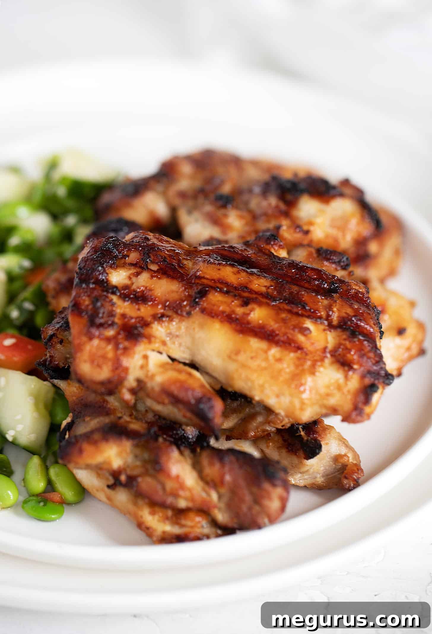 A close-up of grilled miso chicken thighs, beautifully charred and glistening, served with an edamame salad in the background.