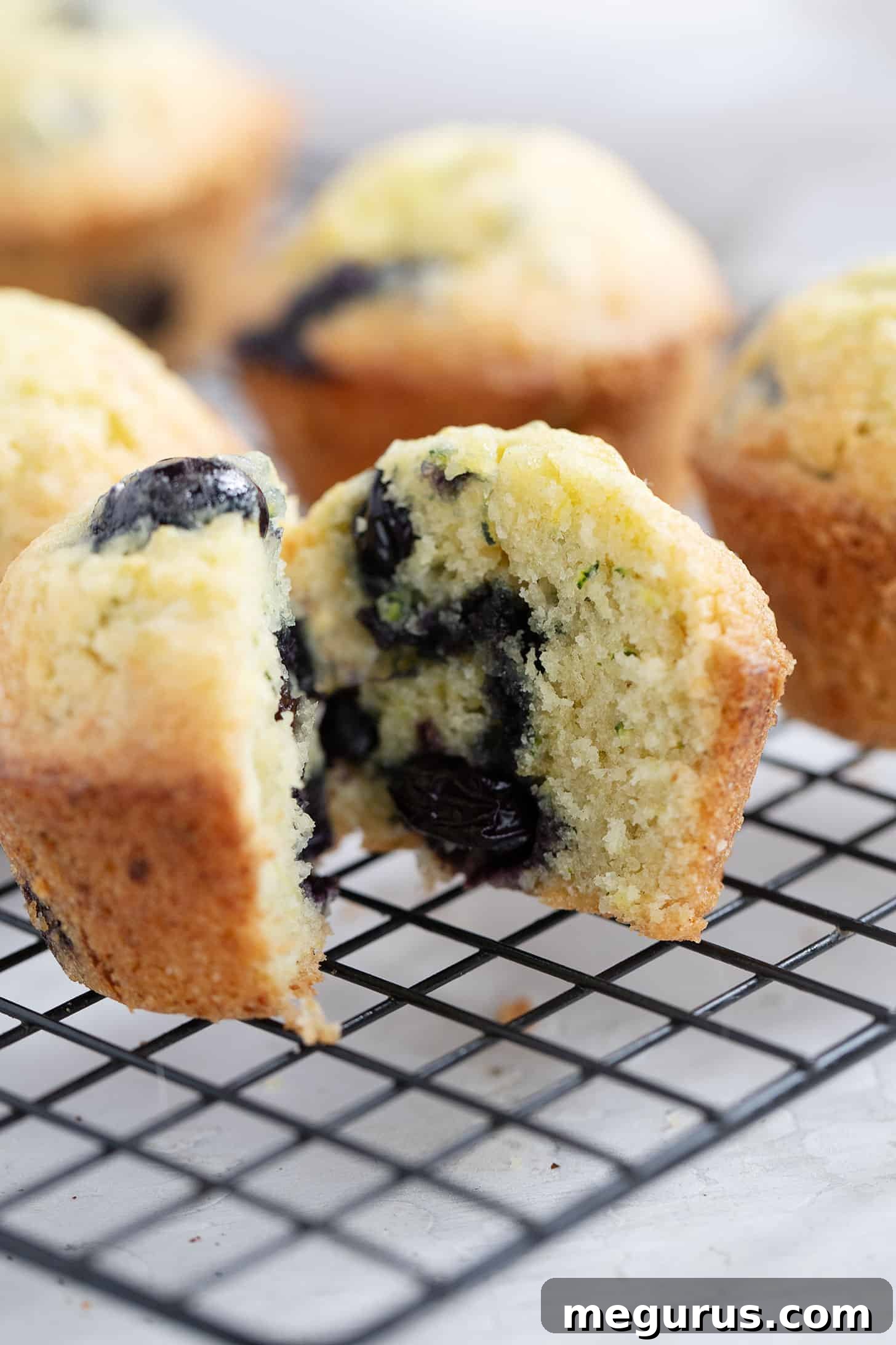 Two blueberry zucchini muffins, one cut in half, showcasing the moist interior and scattered blueberries on a cooling rack.