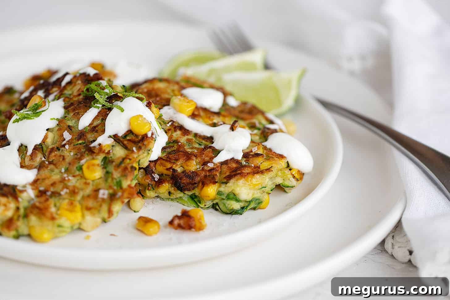 Crispy zucchini corn fritters on a plate drizzled with lime sour cream and garnished with fresh lime zest.