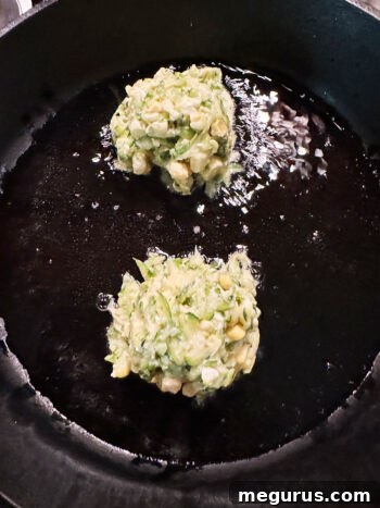 Two fritters in a skillet, still in their initial mound shape before spreading.
