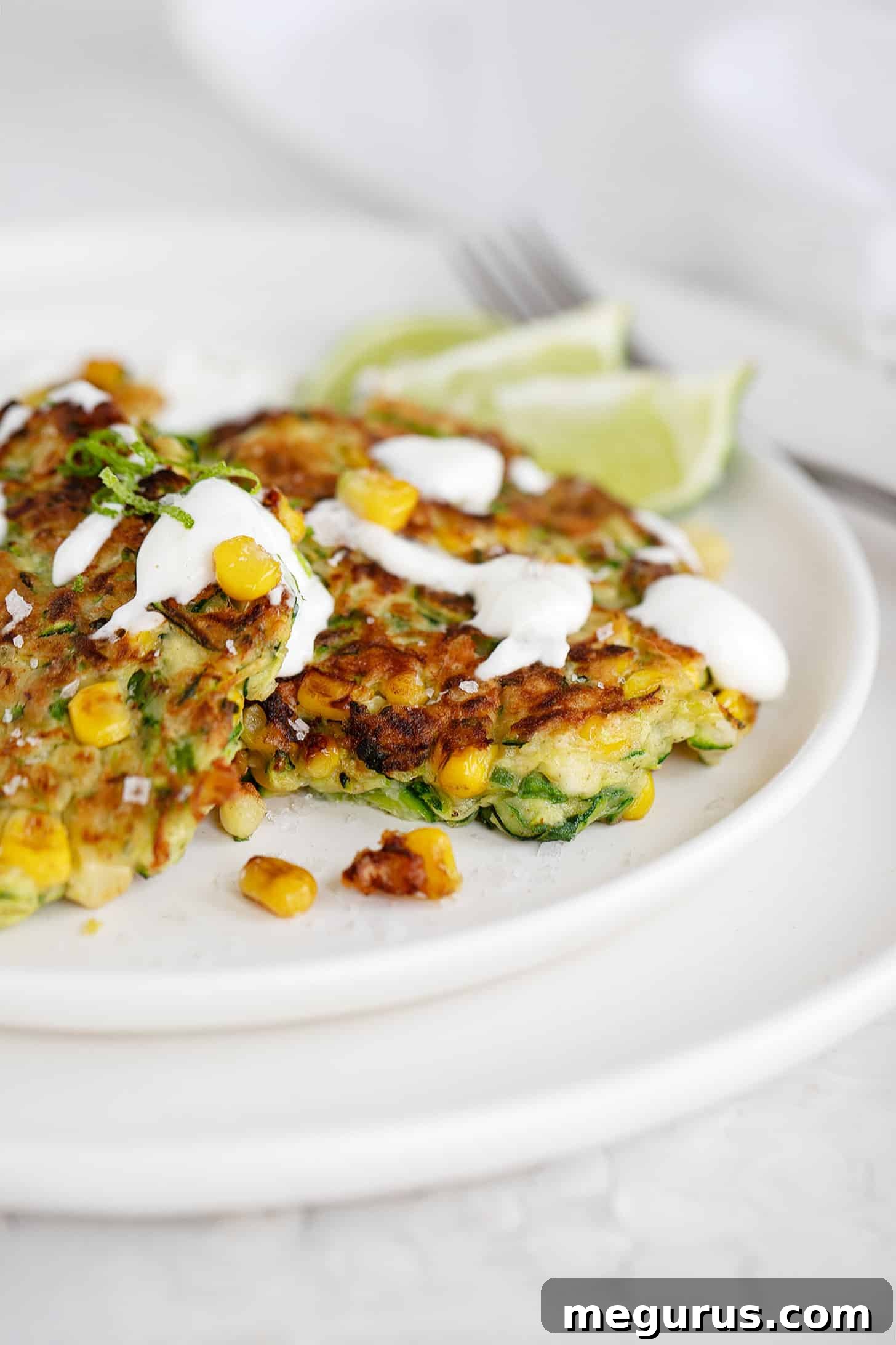 Close-up of zucchini corn fritters on a plate, garnished with lime sour cream, lime zest, and flaky salt.