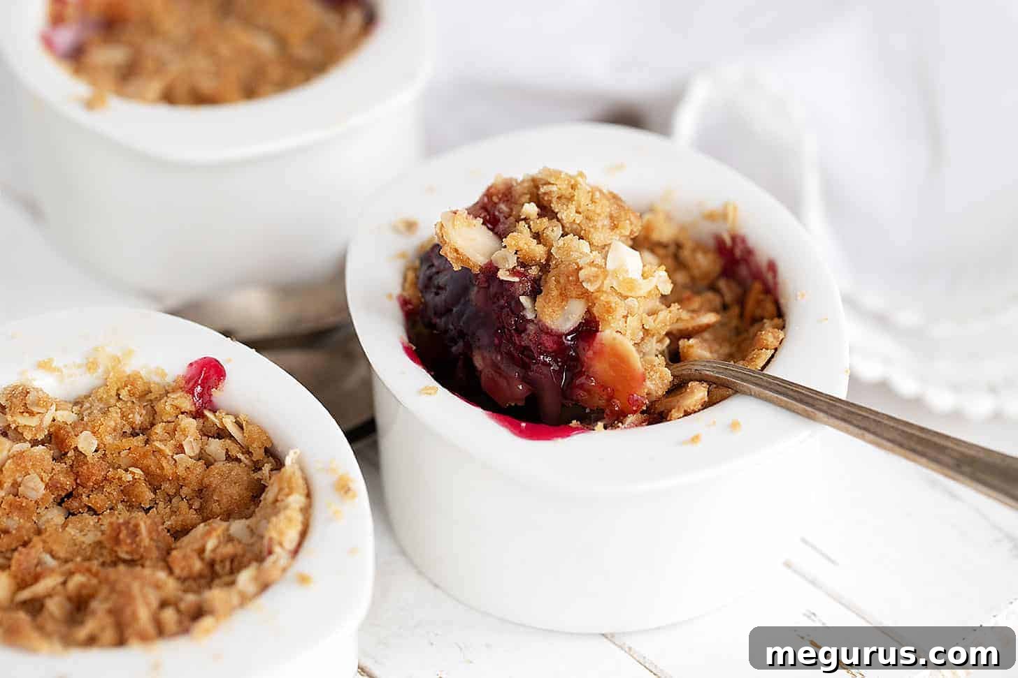 Individual blackberry apple crumbles in ramekins, one with a spoon, highlighting the crispy topping and bubbling fruit filling.
