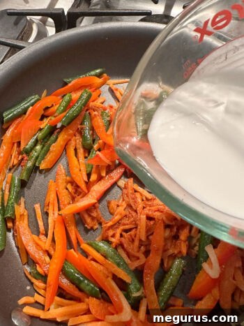 Pouring coconut milk into the skillet with curry paste and vegetables