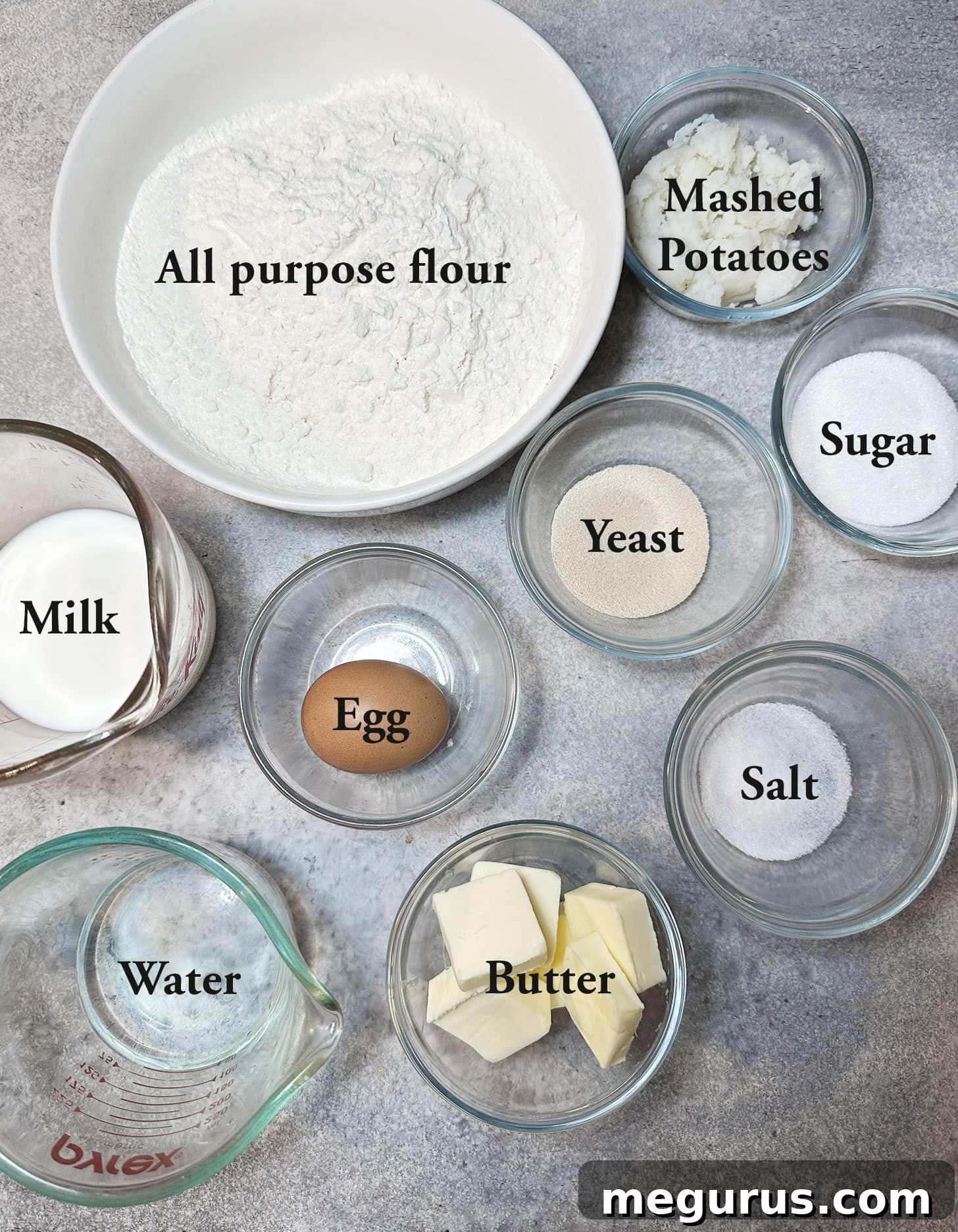 A collection of ingredients laid out on a kitchen counter, including flour, yeast, butter, eggs, milk, sugar, salt, and mashed potatoes, ready for making butterflake rolls.