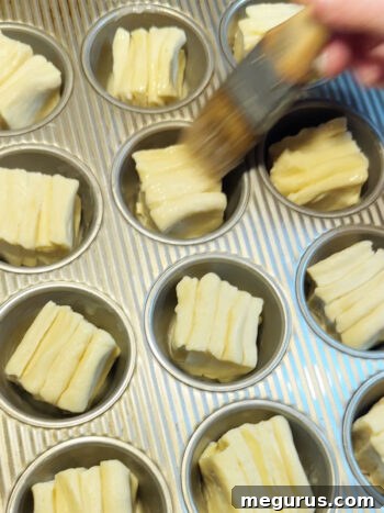 Individual cut rolls placed into a muffin tin and being brushed with butter before the second rise.