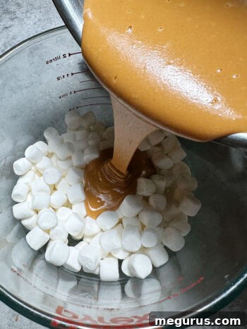 Pouring the slightly cooled peanut butter mixture over a bowl full of mini marshmallows.