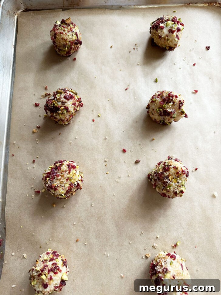 Rolled cookie dough balls coated with cranberry and pistachio mixture on a baking sheet.