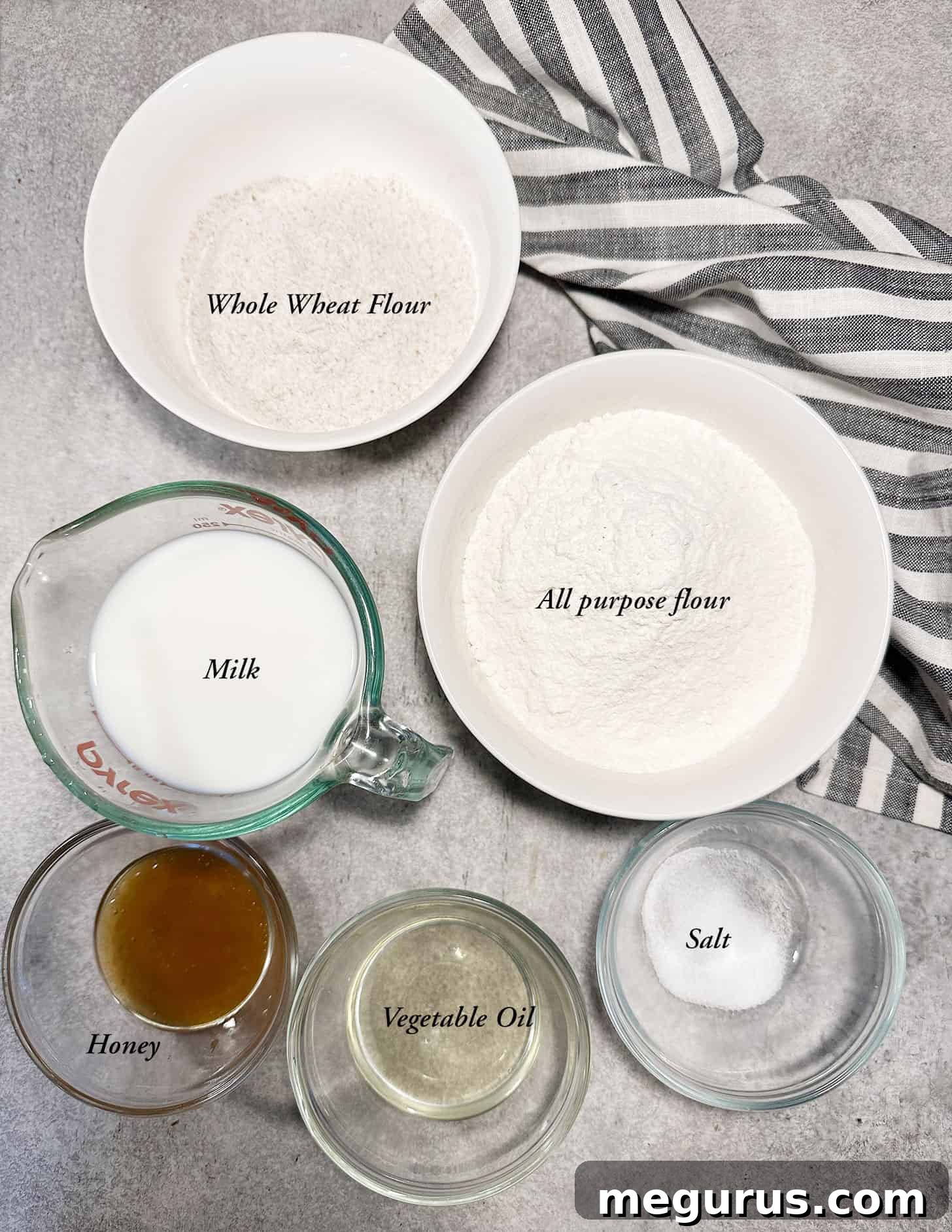 A collection of ingredients laid out, including flour, honey, yeast, and milk, ready for baking honey whole wheat bread.