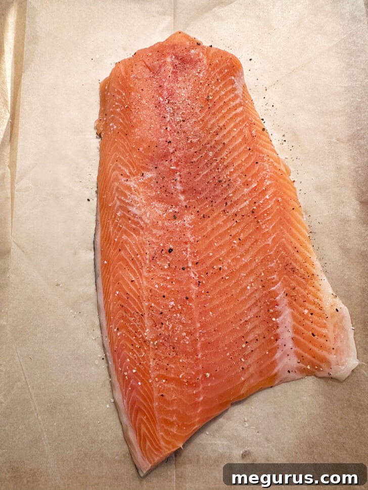 A salmon fillet, skin-side down, is placed on a parchment-lined baking sheet, lightly seasoned with salt and pepper, ready for the topping.