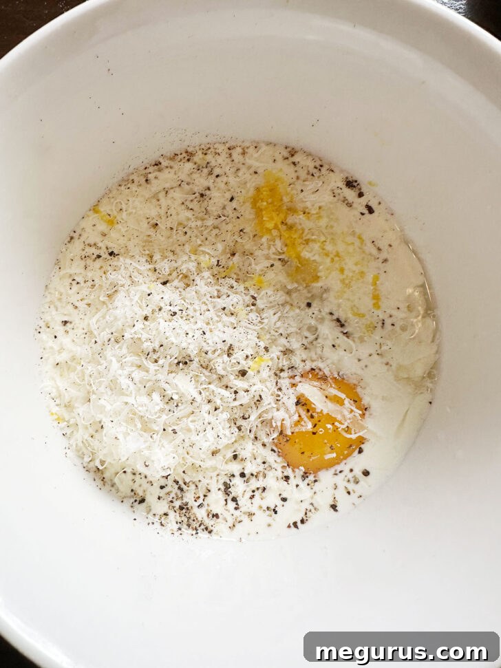 A small bowl containing all the ingredients for the lemon cream sauce: egg yolk, cream, lemon zest, lemon juice, and Parmesan cheese, ready to be whisked.