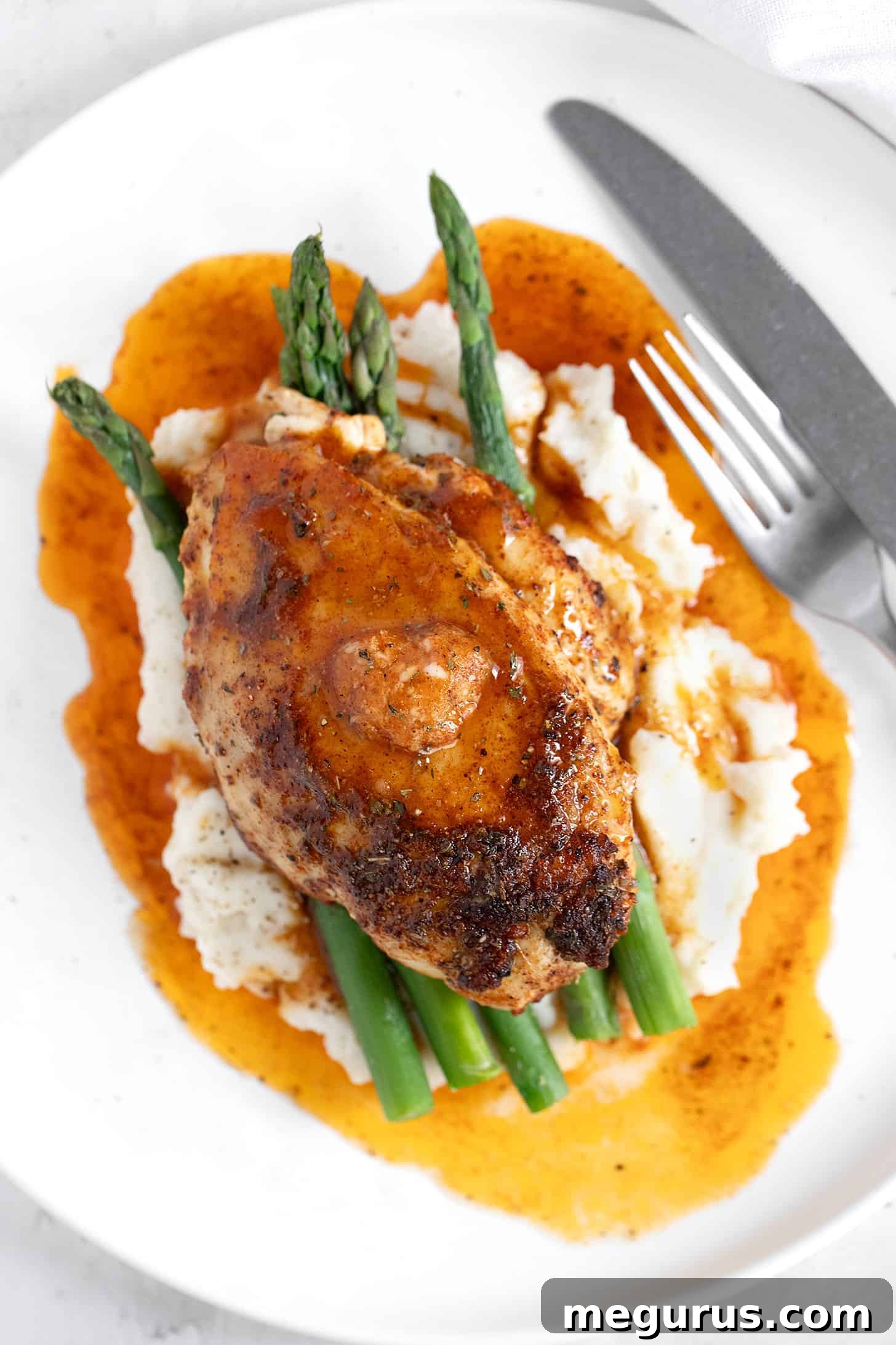 Close-up of a plated Cajun chicken breast with mashed potatoes and asparagus, showing the rich, dark crust and melting honey butter.