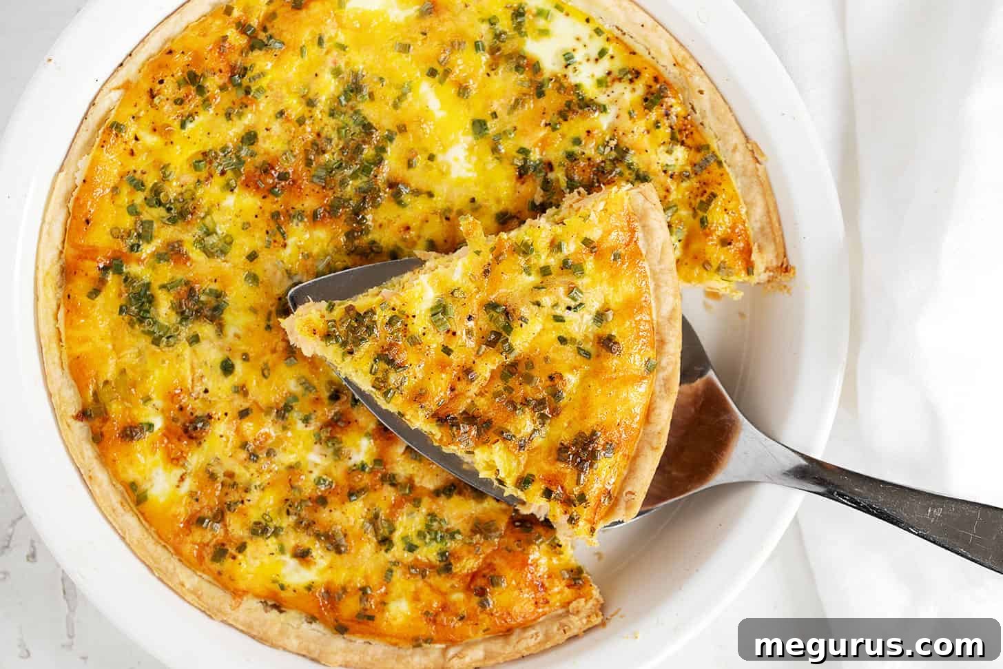 A perfectly baked Salmon Quiche in a pie dish, with a slice already served on a plate, garnished with fresh chives.