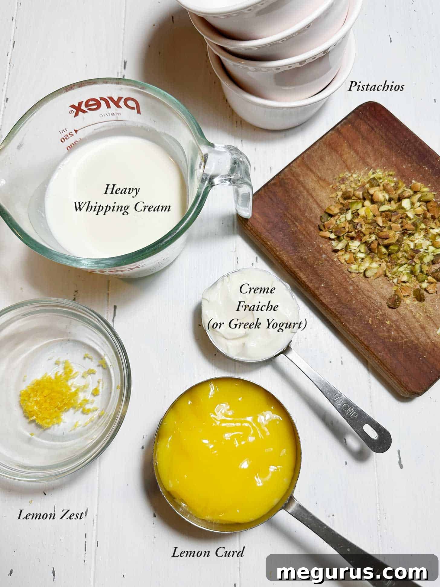 Essential ingredients for lemon curd pudding laid out: lemon curd, crème frâiche, heavy whipping cream, and optional lemon zest and pistachios.