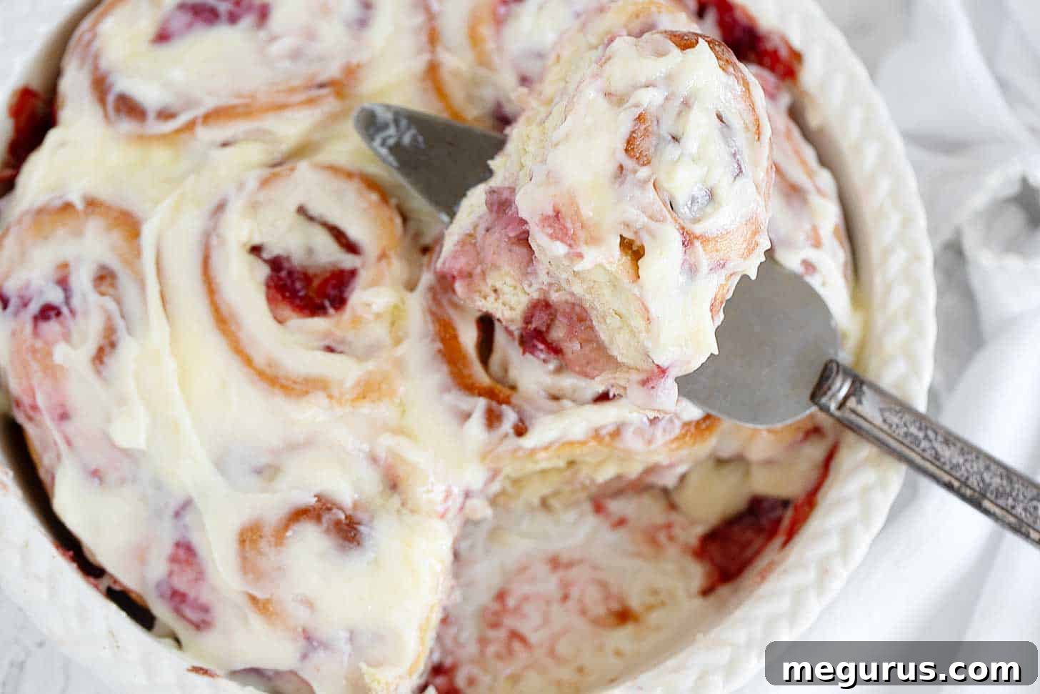Freshly baked strawberry cinnamon rolls in a ceramic baking dish, drizzled with creamy frosting.