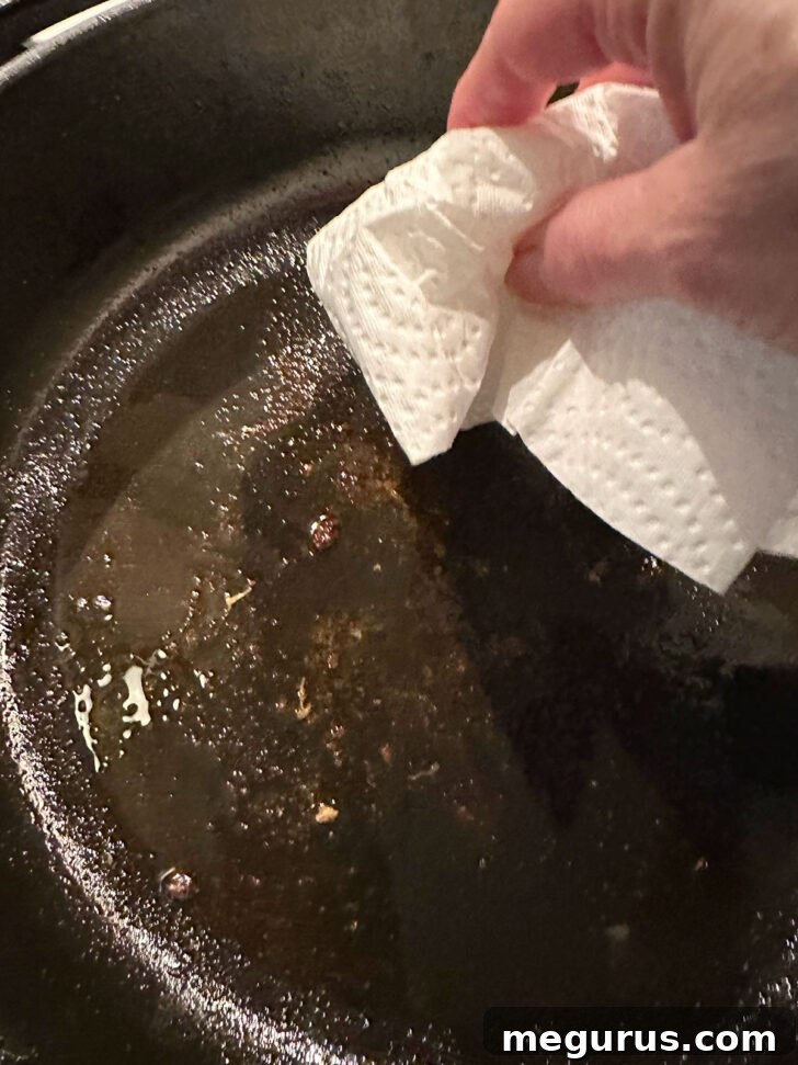 Wiping excess oil from the skillet with a paper towel after searing chicken.