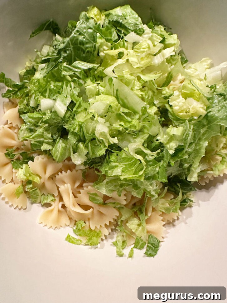 Freshly chopped romaine lettuce being added to the cooked and cooled pasta in a large salad bowl, forming the base of the salad.