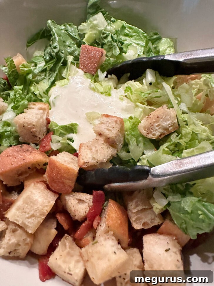 Generous amount of creamy Caesar dressing poured over the assembled salad ingredients in the bowl, ready for a thorough toss.