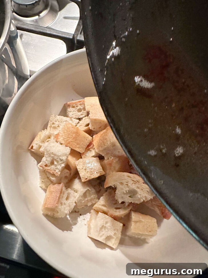 Freshly rendered bacon fat being poured over generously cubed baguette pieces in a mixing bowl, preparing them for seasoning.