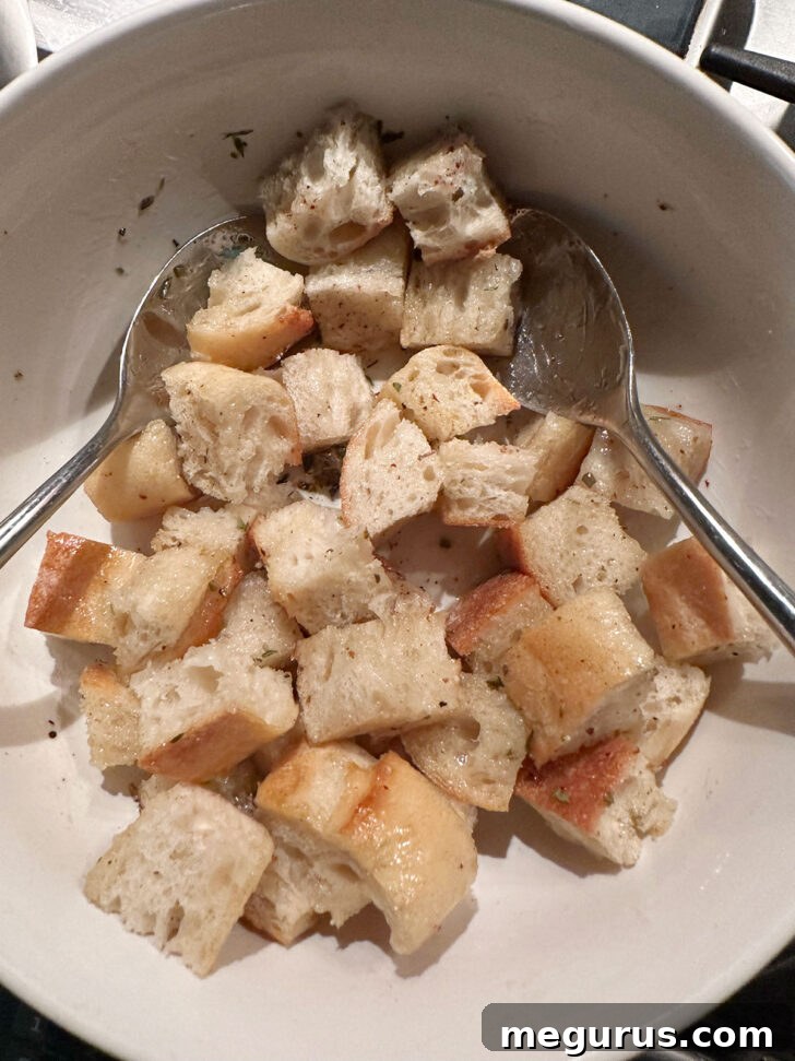 Cubed baguette pieces being thoroughly tossed with bacon fat and a blend of seasonings in a large bowl, ensuring even coating.