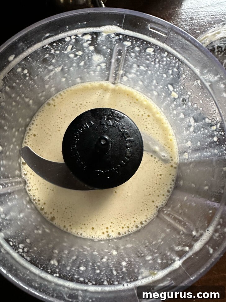 A food processor bowl filled with the initial Caesar dressing ingredients: garlic, salt, vinegar, pasteurized egg, and Dijon mustard.