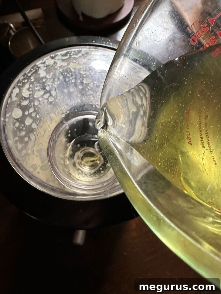 Olive oil being slowly drizzled into the running food processor through the lid's opening, emulsifying with the Caesar dressing base.