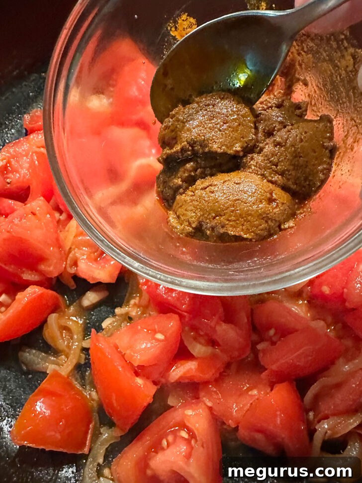 Tikka curry paste being stirred into the skillet with other ingredients.