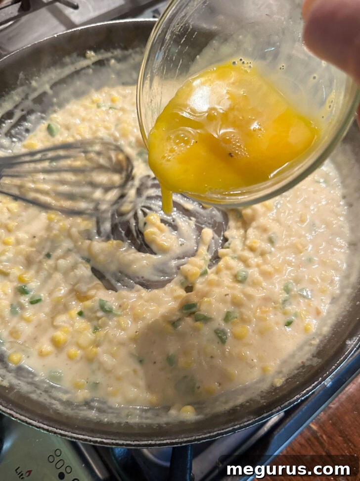 A slightly beaten egg being added to the simmering corn and white sauce mixture in a skillet.