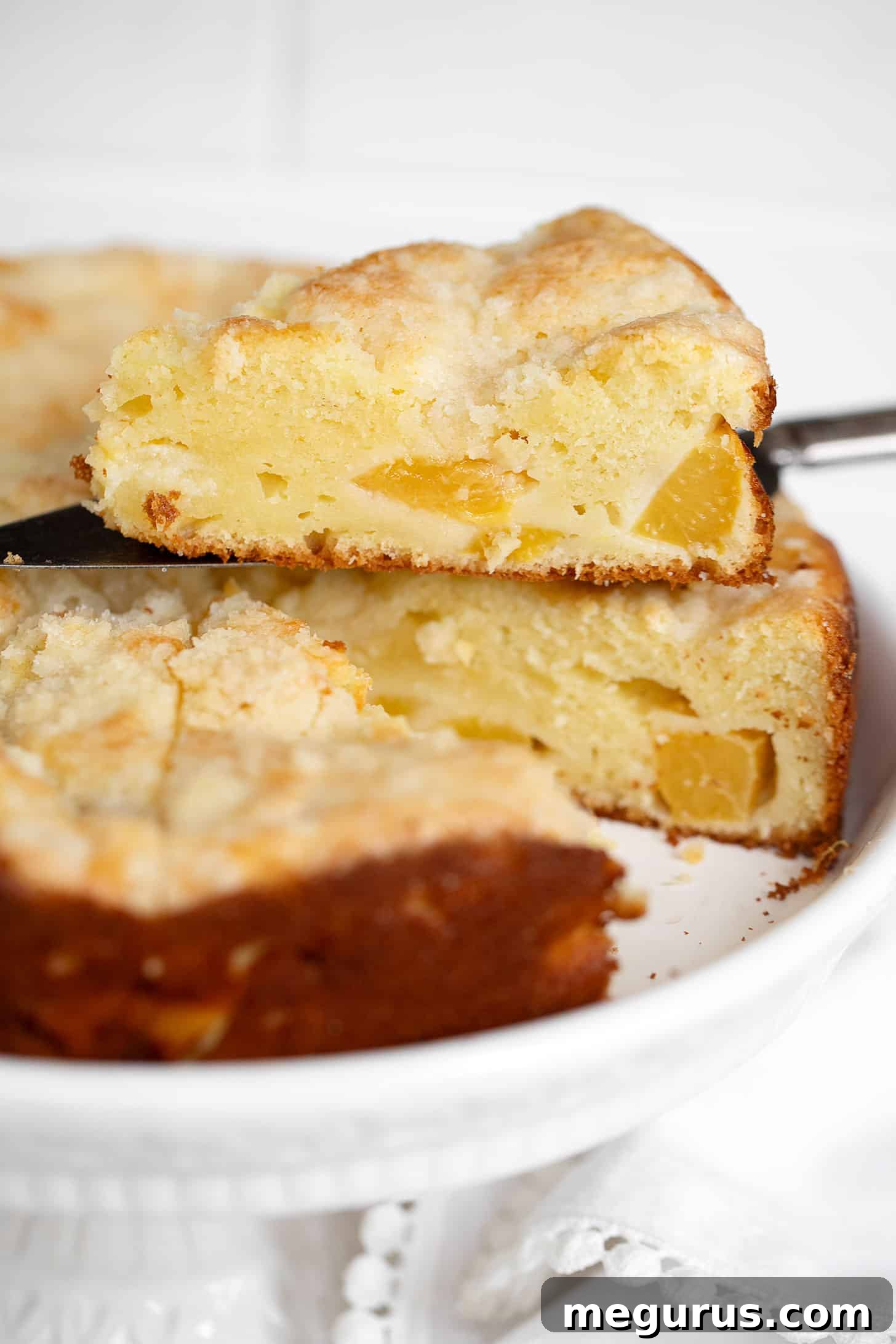 A stunning close-up of a fully baked Peach Cream Cheese Cake, beautifully sliced to reveal its moist interior, abundant peach pieces, and golden crumb topping, presented on an elegant serving plate.