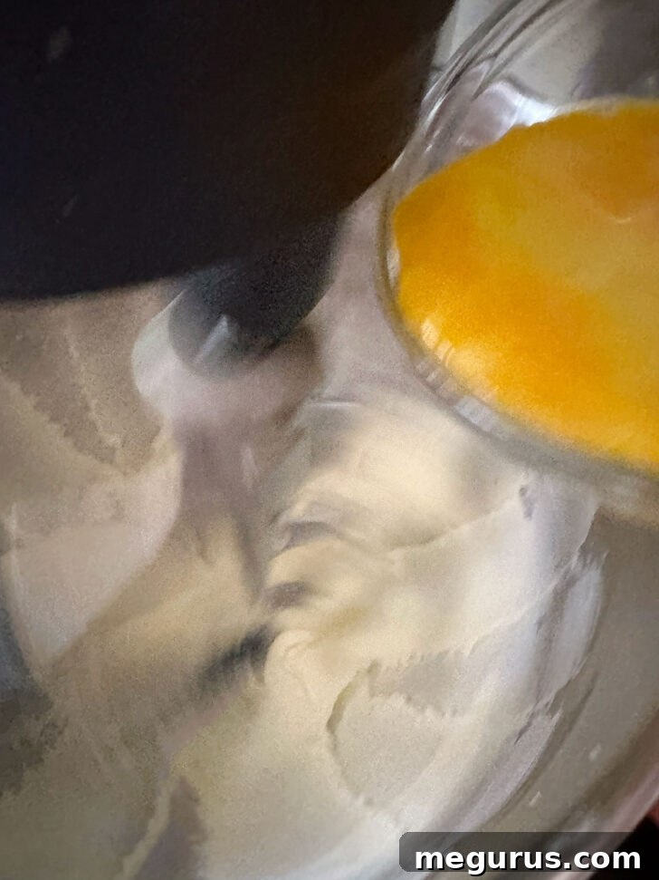 A hand adding a cracked egg to the mixing bowl with creamed butter, sugar, and cream cheese, preparing for the next step of batter preparation.